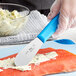 A person's hand with a Choice stainless steel sandwich spreader with a blue handle spreading butter on a piece of fish.