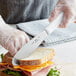 A person using a Choice stainless steel scalloped sandwich spreader to cut a sandwich.