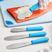 A Choice stainless steel sandwich spreader with a blue handle spreading butter on a piece of fish.