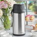 An Acopa stainless steel coffee thermos on a table with a white cup of coffee and a vase of flowers.