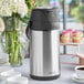 A silver Acopa stainless steel coffee thermos on a table full of coffee cups.