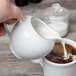 A person using a 10 Strawberry Street Silver Line white porcelain creamer to pour milk into a cup of coffee.