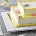 A white rectangular cake board with a cake decorated with yellow and purple flowers.