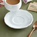 A hand holding a Tuxton bright white demitasse cup of coffee on a saucer with a spoon.