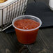 A Dart clear plastic souffle cup filled with red sauce on a table next to a basket of food.