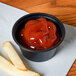 A Dart black plastic souffle cup filled with ketchup on a table with french fries.