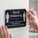 A person holding a Tablecraft plastic "Social Distancing" sign with a symbol on a wall.
