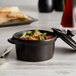 An Acopa black stoneware mini casserole dish with food in it and a lid on a table.