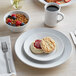 A white Acopa stoneware plate with a biscuit, jam, fruit, and a cup of coffee on it.