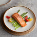 A Acopa stoneware coupe plate with salmon and asparagus on a table with a glass of wine.