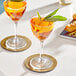 Two Acopa Deco Nick and Nora martini glasses filled with orange liquid and topped with sage leaves on a white table.