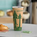 A Kraft EcoChoice paper hot cup with a tree print on it sitting on a table with a croissant and a cup of coffee.