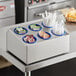 A stainless steel flatware organizer with blue perforated plastic cylinders on a counter with condiments and straws.