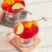 Two glasses of red liquid with lime slices and orange peels with a red bamboo knot skewer in one glass.