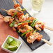 A grilled shrimp and vegetable skewer on a black plate with a Bamboo by EcoChoice bamboo paddle food pick.