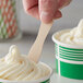 A hand holding a Choice 3" Eco-Friendly Unwrapped Wooden Taster Spoon over a bowl of ice cream.