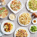 A table with plates of pasta, salad, and other foods including bowls of Barilla whole grain penne with vegetables and bacon and cheese.