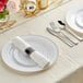A white table setting with Visions silver banded plastic dinnerware and rolled hammered flatware on a white plate with a napkin.