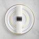 A white Visions plastic plate with a black and gold banded rim with rolled Classic flatware on it.