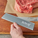 A hand holding a Schraf knife with a gray polypropylene blade guard cutting a piece of meat on a counter.
