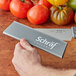 A hand holding a Schraf knife with a gray polypropylene blade guard cutting a tomato.