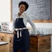 A woman wearing a navy blue bib apron with natural webbing accents standing in front of a counter.
