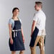 A man and woman wearing navy blue Choice bib aprons with natural webbing accents.