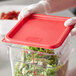 A person wearing gloves is holding a Carlisle food storage container with a red lid filled with leafy greens.