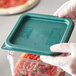 A gloved hand holding a Carlisle green square polypropylene food storage container lid over a container of food.