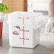 A person using a measuring cup to pour ingredients into a white Carlisle food storage container with red lettering.
