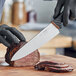 A person in black gloves uses a Schraf 8" Chef Knife with a brown handle to cut a piece of meat.