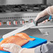 A hand holding a Schraf serrated slicing knife with a blue TPRgrip handle cutting salmon on a blue cutting board.