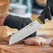 A person with black gloved hands uses a Schraf Santoku knife to cut meat on a cutting board.