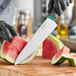 A person in gloves uses a Schraf chef knife with a green handle to cut a watermelon on a wooden surface.
