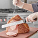 A person using a Schraf serrated slicing knife with a brown handle to cut a piece of meat.