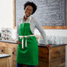 A smiling woman wearing a Choice Kelly Green Poly-Cotton Bib Apron standing in front of a counter.