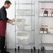 A man in an apron and gloves standing next to a Baker's Mark metal shelving unit with white plastic ingredient bins.