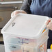 A woman in a blue apron holding a Vigor white square polypropylene food storage container with a plastic lid.