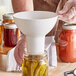 A person using a white Vigor wide mouth canning funnel to pour pickles into a jar.