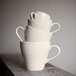 A stack of white Tuxton cappuccino mugs with a curved handle.
