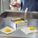 Stainless steel square egg ring cooking a fried egg in a metal pan.