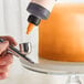 A finger holding Chefmaster Sunset Orange Airbrush Color over a cake.