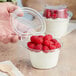 A hand holding a clear plastic parfait cup with a bowl of yogurt and raspberries.