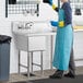 A person in a blue apron and gloves cleaning a Regency stainless steel commercial sink in a professional kitchen.
