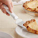 A hand using a Choice pie server to cut a slice of pie on a plate.