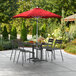 A red Lancaster Table & Seating umbrella with a white pole over a table and chairs on a patio.