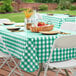 A picnic table with a green and white checkered vinyl tablecloth and a basket of fruit.