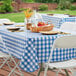 A picnic table with a Choice royal blue and white checkered vinyl tablecloth and a basket of fruit.