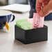 A hand putting a green and pink packet of sugar in a black stainless steel rectangular sugar caddy.