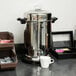 A Hamilton Beach stainless steel coffee urn on a table with a white mug next to it.
